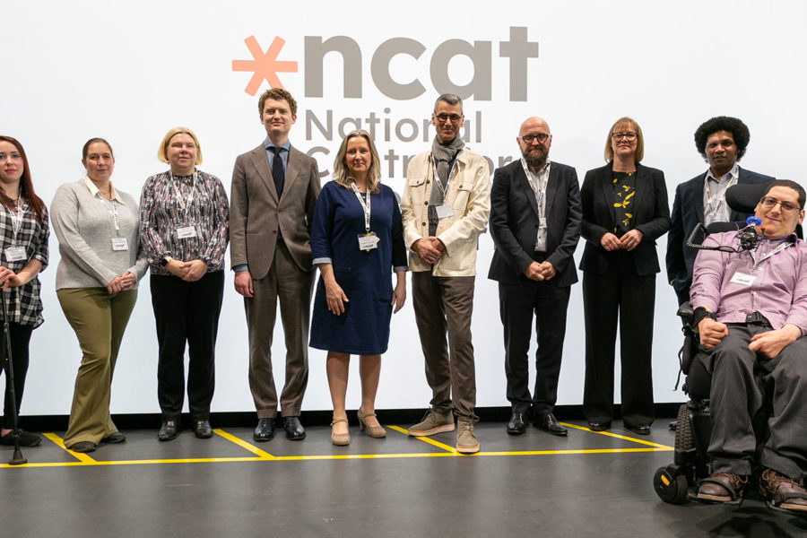 A mixed group of people standing, and one person in a wheelchair, in front of a large display screen which showing the ncat logo. They are all smiling.