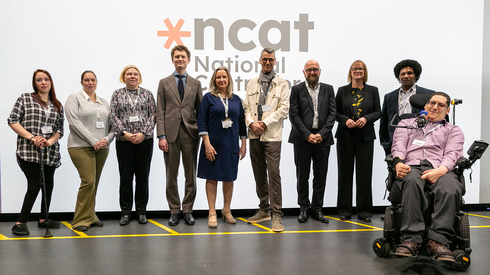 A mixed group of people standing, and one person in a wheelchair, in front of a large display screen which showing the ncat logo. They are all smiling.