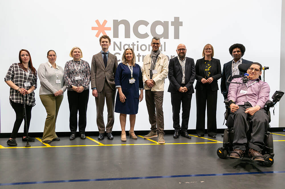 A mixed group of people standing, and one person in a wheelchair, in front of a large display screen which showing the ncat logo. They are all smiling.