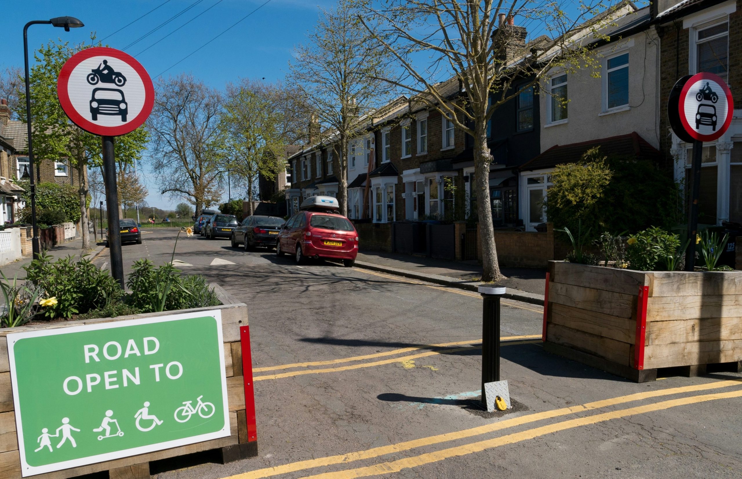 street scene showing parked cars on side of street, yellow lines painted on road, trees, sign prohibiting cars and motorbikes, green sign saying 'Road Open to' and then symbols for pedestrians, skaters, wheelchair users, and cyclists. Two large wooden plant boxes placed on either side of the road.