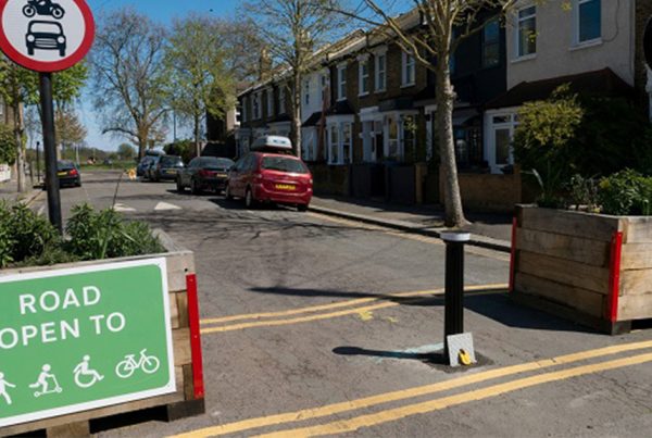 street scene showing parked cars on side of street, yellow lines painted on road, trees, sign prohibiting cars and motorbikes, green sign saying 'Road Open to' and then symbols for pedestrians, skaters, wheelchair users, and cyclists. Two large wooden plant boxes placed on either side of the road.