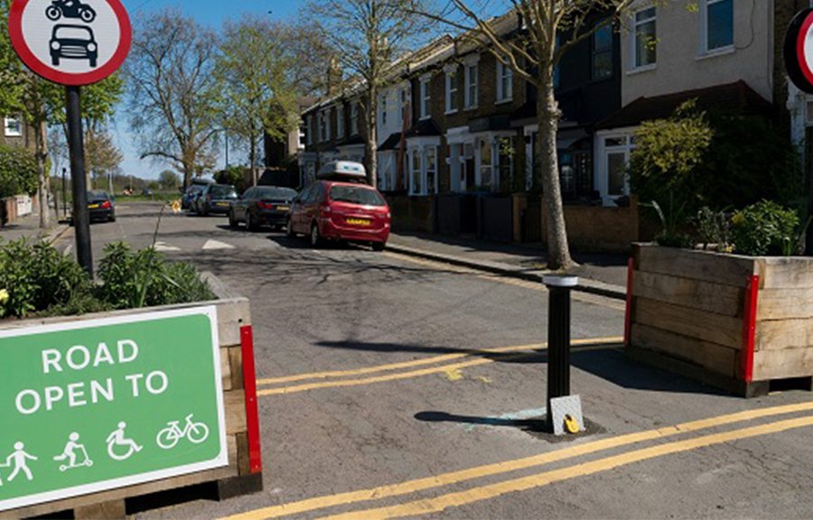 street scene showing parked cars on side of street, yellow lines painted on road, trees, sign prohibiting cars and motorbikes, green sign saying 'Road Open to' and then symbols for pedestrians, skaters, wheelchair users, and cyclists. Two large wooden plant boxes placed on either side of the road.