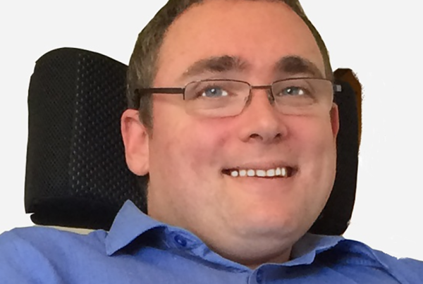 smiling headshot photo of ncat Board member Damian Bridgeman, wearing blue shirt and glasses sitting in a chair
