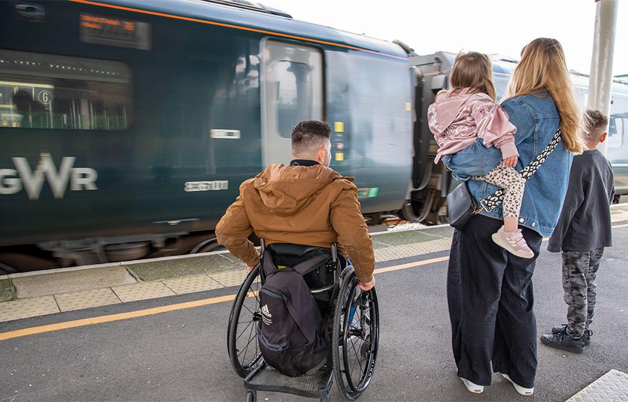 Family with their backs to the camera, waiting on a railway platform with train in front of them. One adult in a wheelchair, one adult holding a small cild, with another child alongside.