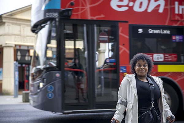 woman standing facing camera holding mobility aids. Bus in background in front of Coventry Pool Meadow Bus Station