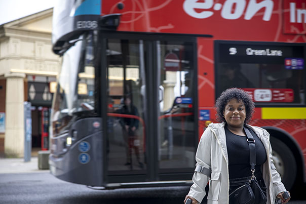 woman standing facing camera holding mobility aids. Bus in background in front of Coventry Pool Meadow Bus Station