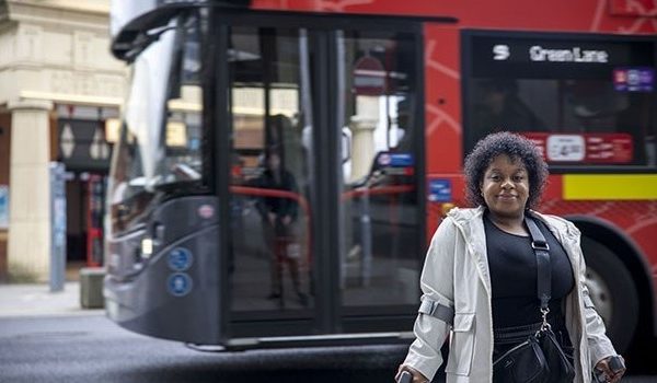 woman using mobility aids facing camera standing in front of a double decker bus and Coventry Pool Meadow bus station