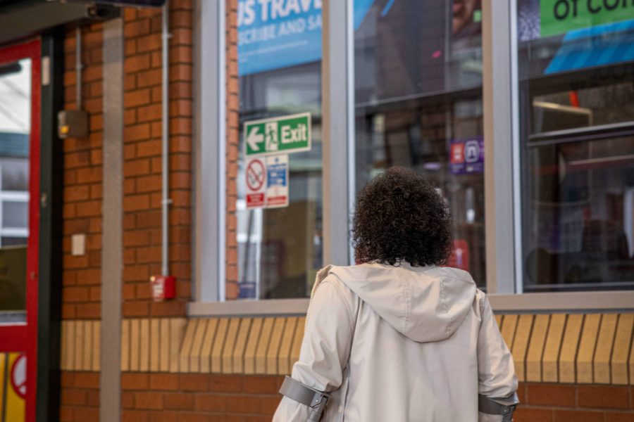 Woman in white coat walking using mobility aids in bus station. Double decker bus can be seen in background through windows. Screen in top left of image for bus times