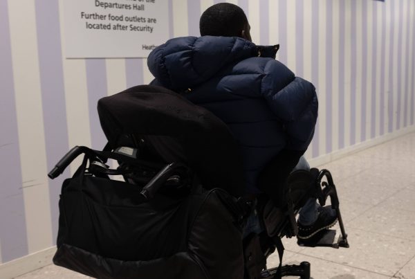 Male wheelchair user facing away from the camera is in front of a Passenger Information sign which states that 'Food and drink is available at The Queen's Arms at the opposite end of the departure hall.' Above this writing are symbols of a sandwich and drinks. Further on the right of the picture is another sign which says 'Assistance' and has an arrow pointing to the right.
