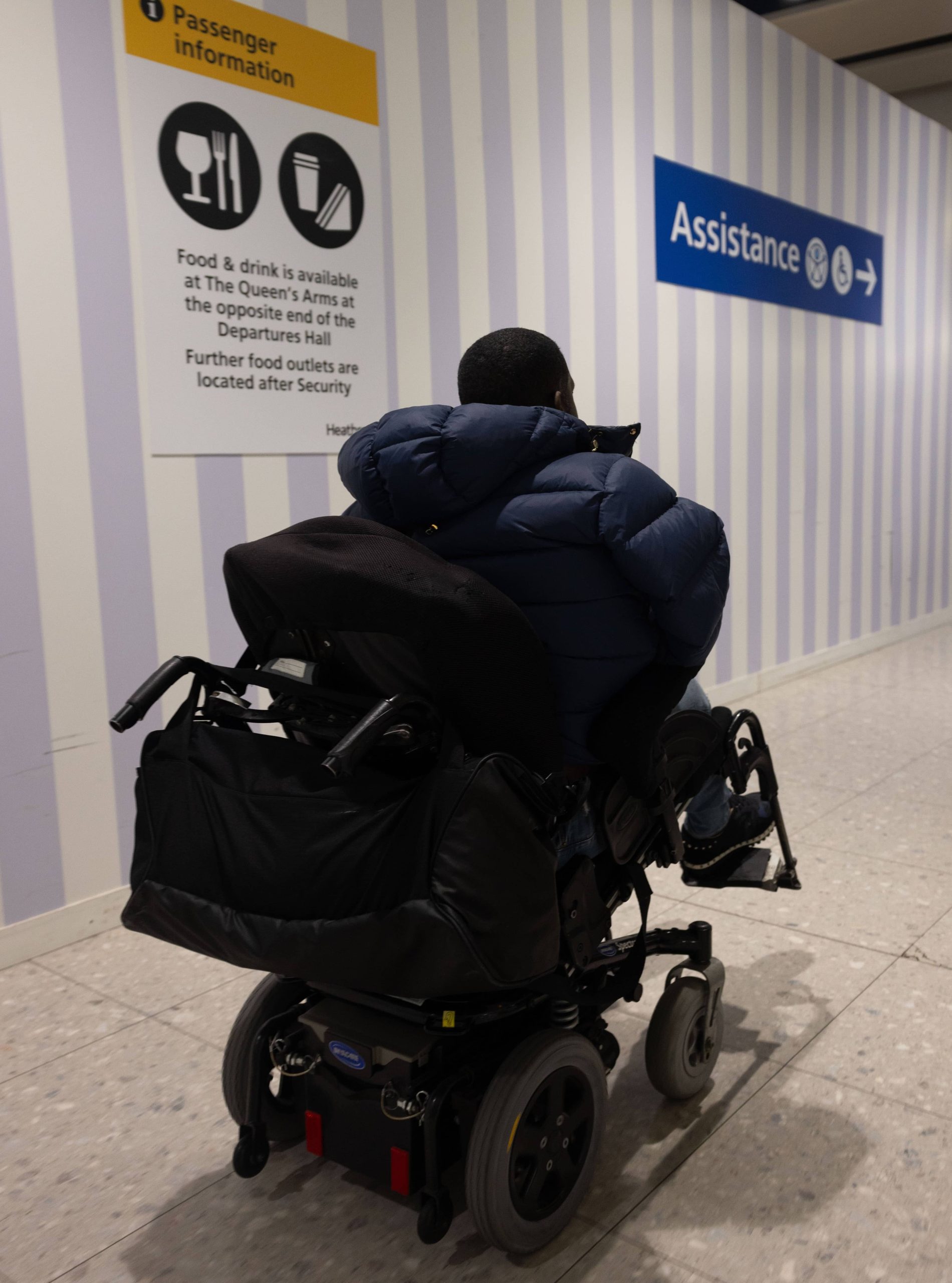 Male wheelchair user facing away from the camera is in front of a Passenger Information sign which states that 'Food and drink is available at The Queen's Arms at the opposite end of the departure hall.' Above this writing are symbols of a sandwich and drinks. Further on the right of the picture is another sign which says 'Assistance' and has an arrow pointing to the right.