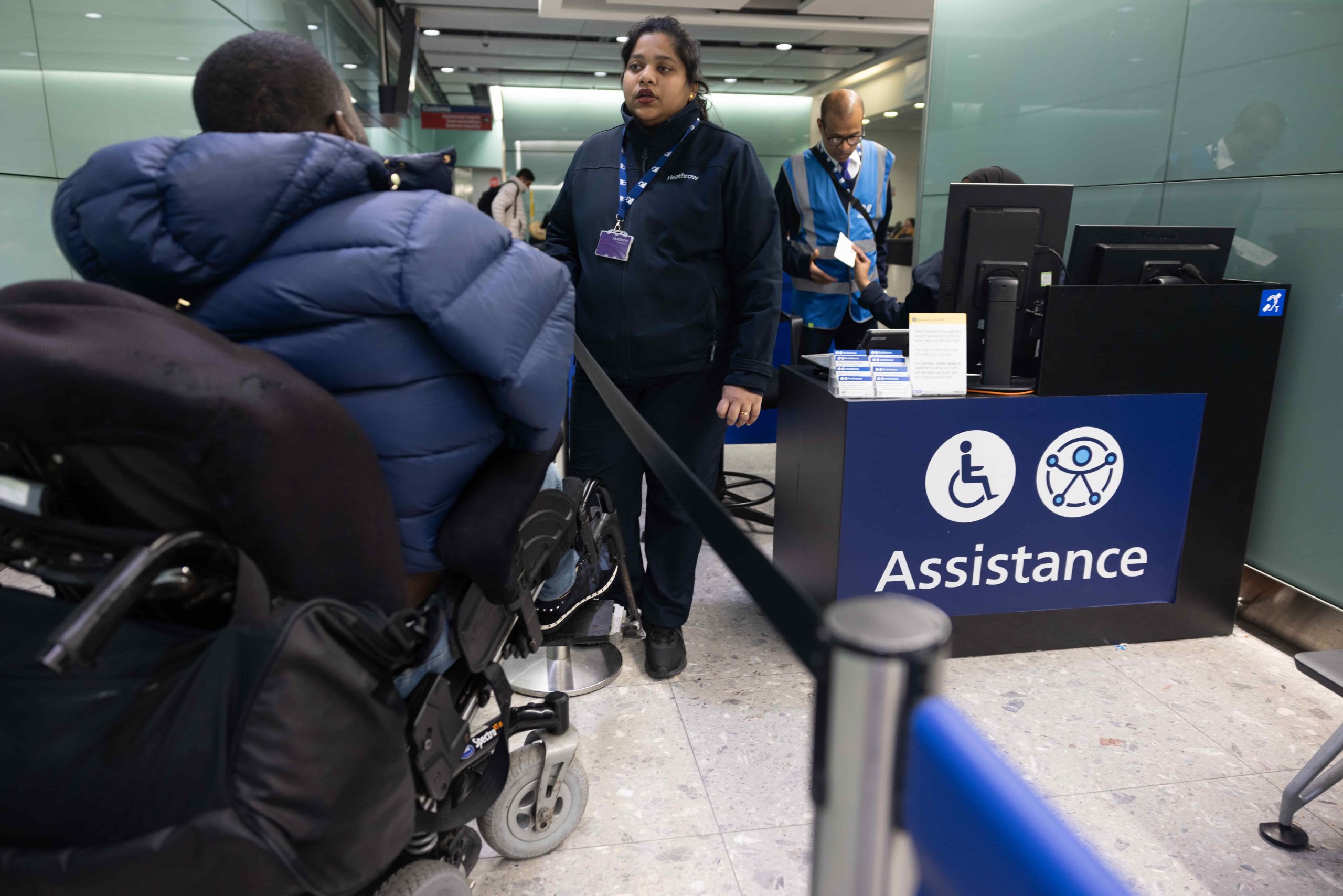 Male wheelchair user with his back to the camera faces a female airport staff member. There is another male staff members in the background. On the right of the image is a sign which reads 'Assistance' and has a wheelchair symbol on it.
