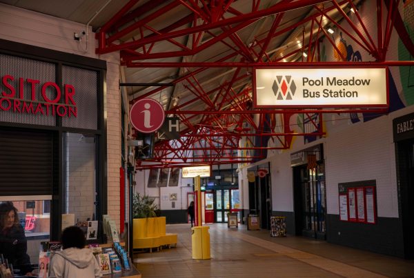 woman standing at visitor information on left of photo. On the right of the image is a open space in Pool Meadow Bus Station. Several signs are hanging from ceiling showing bus times and destinations