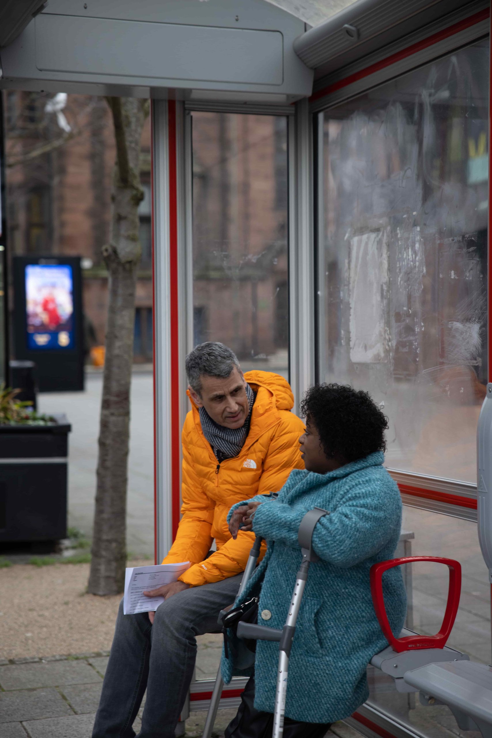 Man in orange coat talks to women in blue coat at bus shelter. A tree and flower pot are in the background of the image.