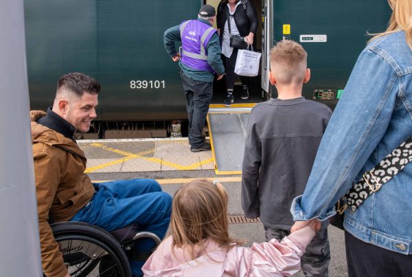 Male wheelchair user on left looks at his young daughter (middle) while waiting with his family (son and female partner) to board the train. In the background is a train operative who has positioned a ramp up to the carriage. He is talking to a passenger before they exit the train.