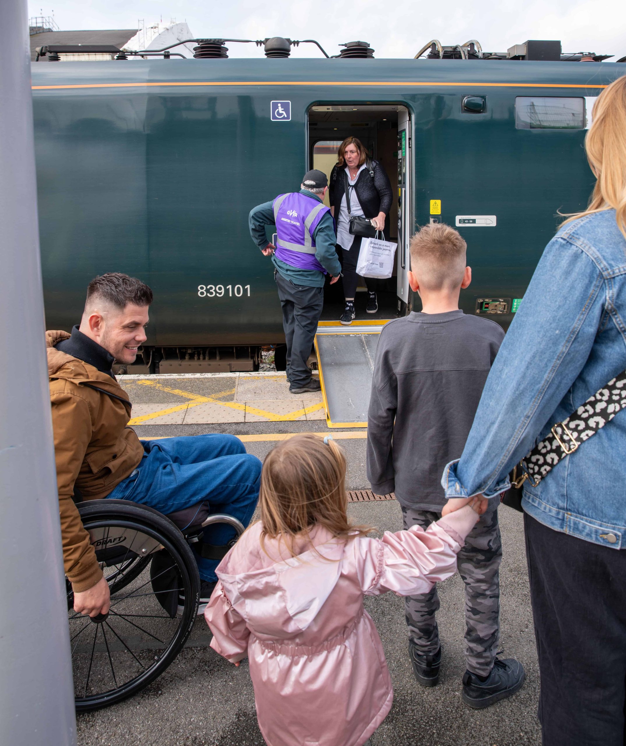 Male wheelchair user on left looks at his young daughter (middle) while waiting with his family (son and female partner) to board the train. In the background is a train operative who has positioned a ramp up to the carriage. He is talking to a passenger before they exit the train.