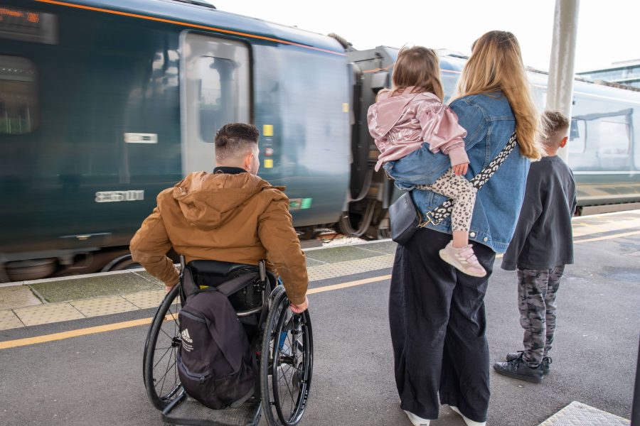 male wheelchair user and his partner and two children wait on train platform in from of a train