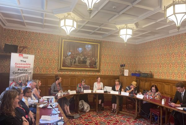 Eleven people sitting around a U-shaped set of tables in a room in the House of Lords. An old painting is on the wall behind them and a banner reading 'The Accessible Transport Policy Commission' is behind the people on the left.
