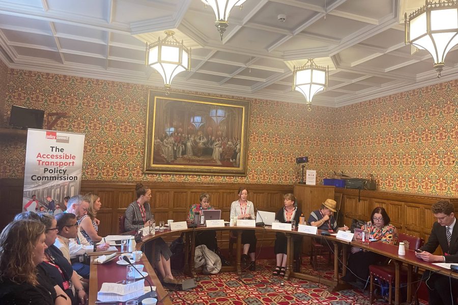 Eleven people sitting around a U-shaped set of tables in a room in the House of Lords. An old painting is on the wall behind them and a banner reading 'The Accessible Transport Policy Commission' is behind the people on the left.