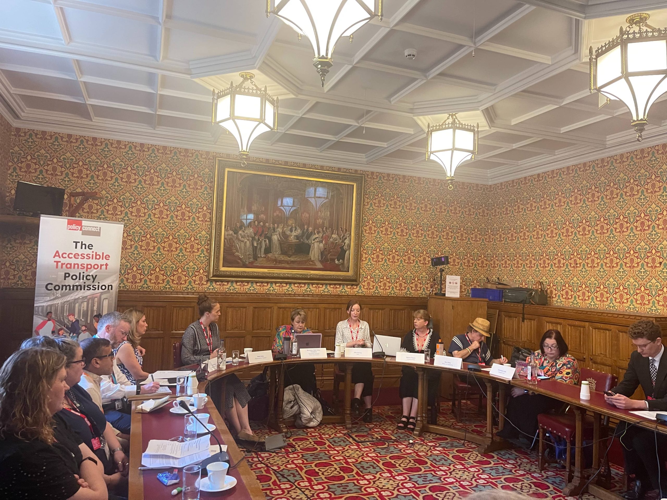 Eleven people sitting around a U-shaped set of tables in a room in the House of Lords. An old painting is on the wall behind them and a banner reading 'The Accessible Transport Policy Commission' is behind the people on the left.