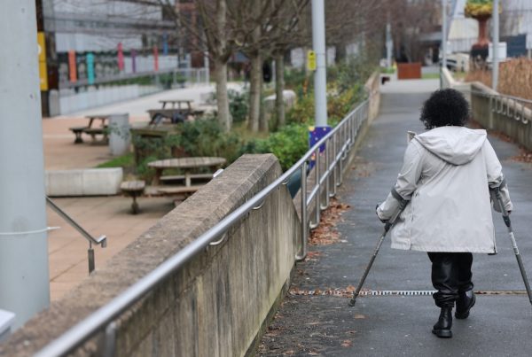 Woman walking using mobility aids, with her back to the camera - in a raincoat, walking down a pathway, adjacent to outdoor seating spaces - urban landscape