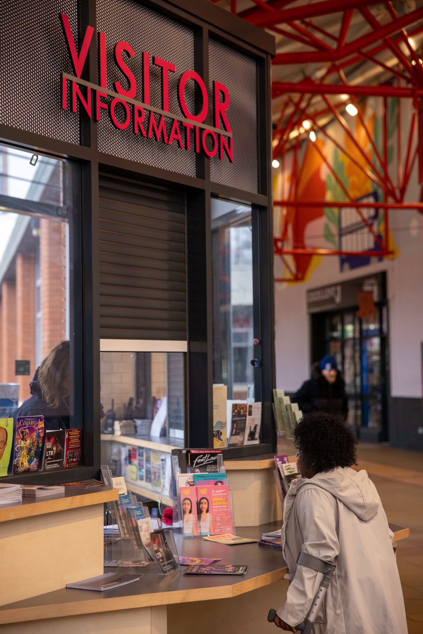 Woman using mobility aids is standing at the Visitor Information desk in Coventry Pool Meadow Bus Station. There are lots of leaflets on the desk and a staff member is slightly obscured by the leaflets on the other side of the desk.