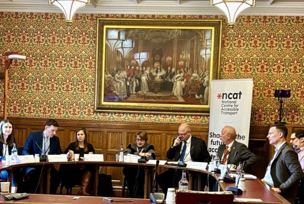Baroness Tanni Grey-Thompson (centre) chairing Accessible Transport Policy Commission Event. There is a semi circle of tables with eight people (six male, two female) sitting behind them facing the camera. A standing poster sits behind them reading 'National Centre for Accessible Transport'. They are in the House of Lords and a very large old painting hangs on the wall behind them.