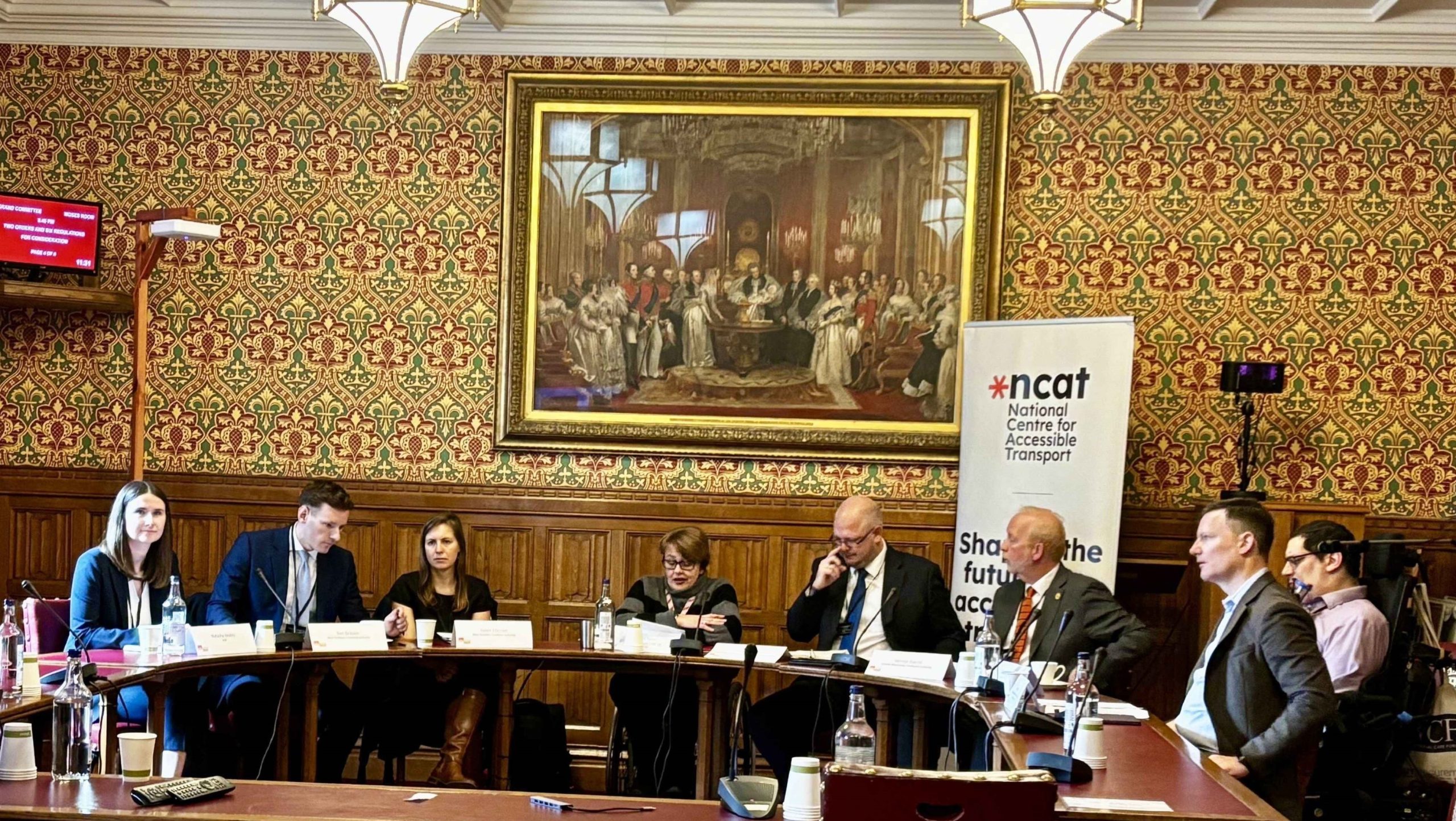 Baroness Tanni Grey-Thompson (centre) chairing Accessible Transport Policy Commission Event. There is a semi circle of tables with eight people (six male, two female) sitting behind them facing the camera. A standing poster sits behind them reading 'National Centre for Accessible Transport'. They are in the House of Lords and a very large old painting hangs on the wall behind them.