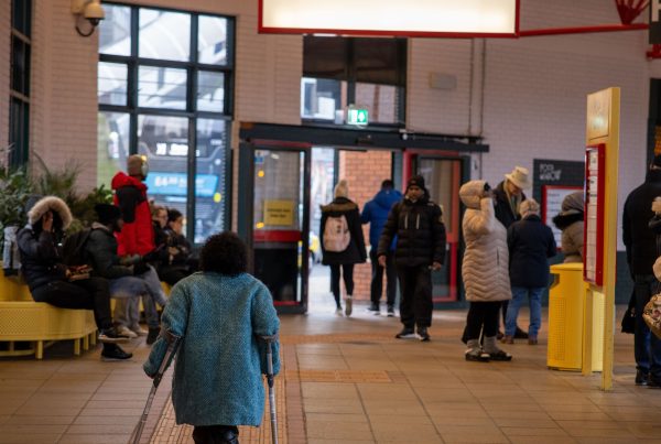 woman in blue coat walking away from camera using mobility aids. She is in Coventry Pool Meadow Bus station. About a dozen members of the public in winter coats are sitting in the station, reading a timetable or walking towards the exit.