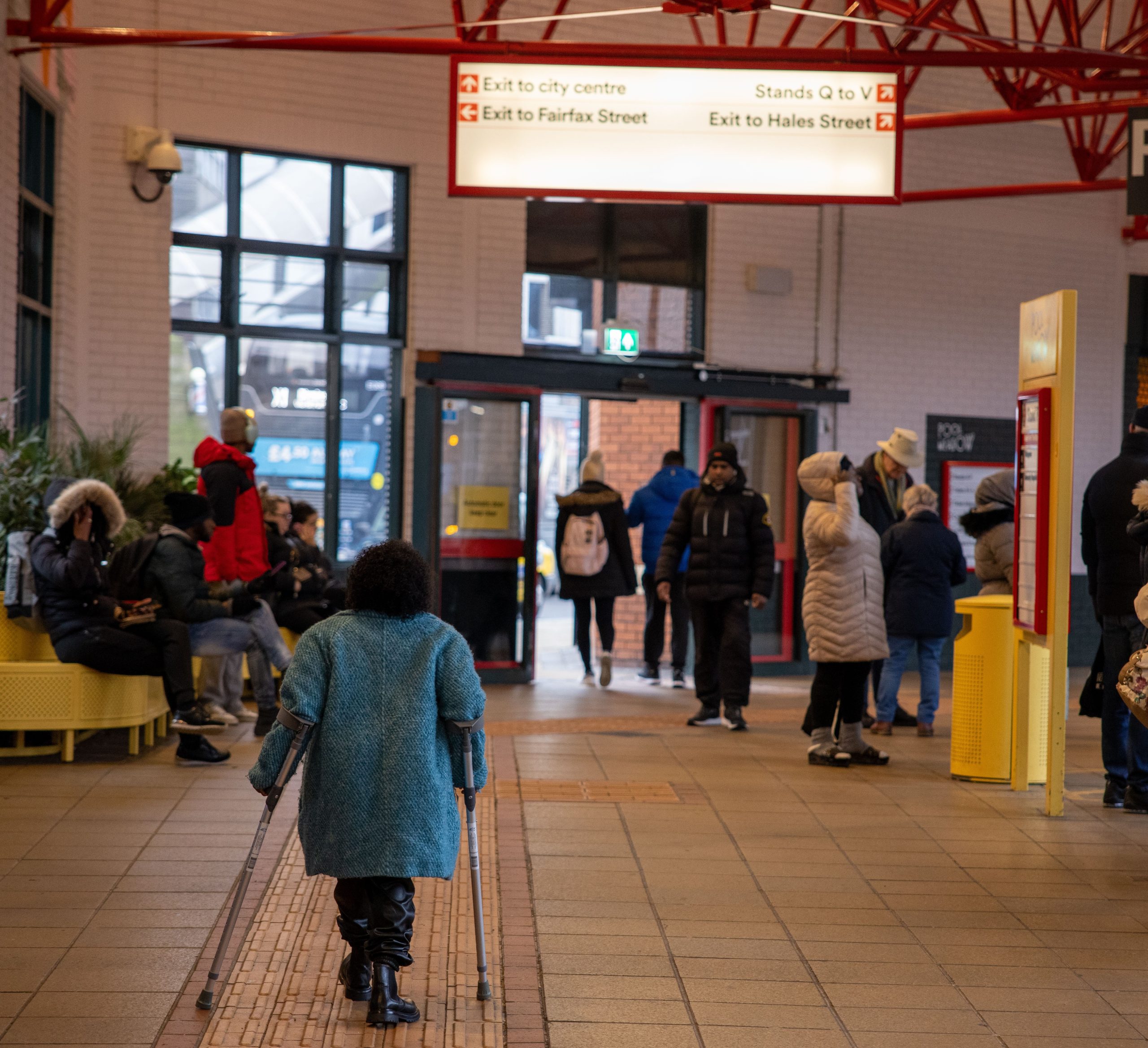 woman in blue coat walking away from camera using mobility aids. She is in Coventry Pool Meadow Bus station. About a dozen members of the public in winter coats are sitting in the station, reading a timetable or walking towards the exit.