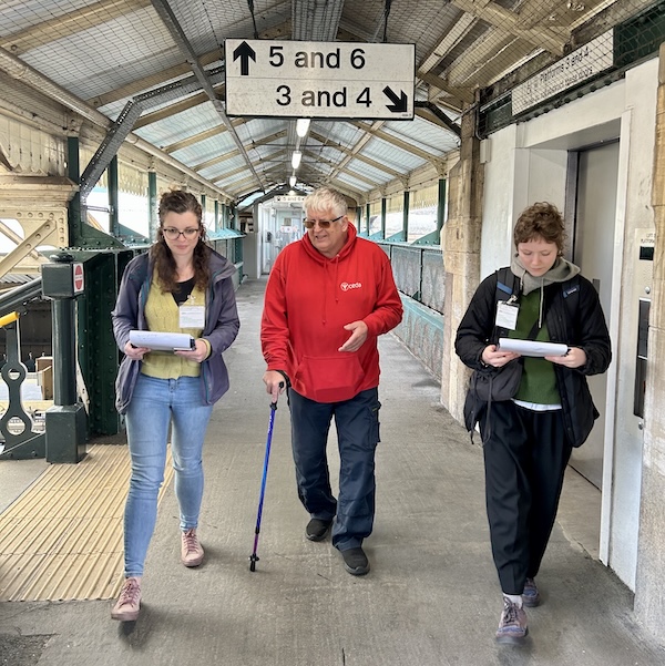 Three people walking on a train platform. The man in the centre carries a walking cane and wears glasses. The two women either side carry clipboards and wear identification badges