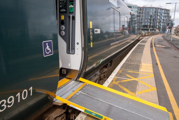 Train carriage waiting at platform. Door is open with assistance ramp in place to platform. Person in wheelchair on blue background symbol is pictured on the train next to the door.