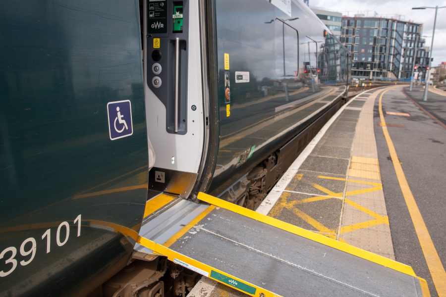 Train carriage waiting at platform. Door is open with assistance ramp in place to platform. Person in wheelchair on blue background symbol is pictured on the train next to the door.