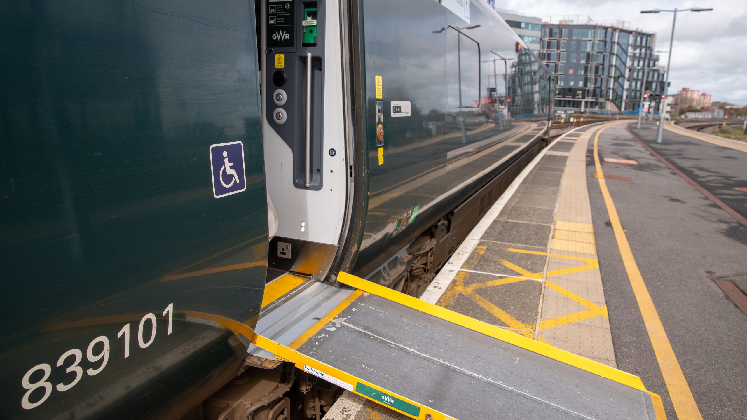 Train carriage waiting at platform. Door is open with assistance ramp in place to platform. Person in wheelchair on blue background symbol is pictured on the train next to the door.