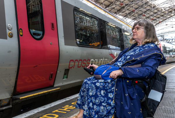 Female wheelchair user in blue dress waits on train platform behind tactile paving. Train, tracks and station to her left in the image.