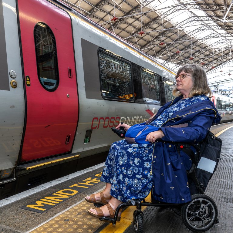 Female wheelchair user in blue dress waits on train platform behind tactile paving. Train, tracks and station to her left in the image.