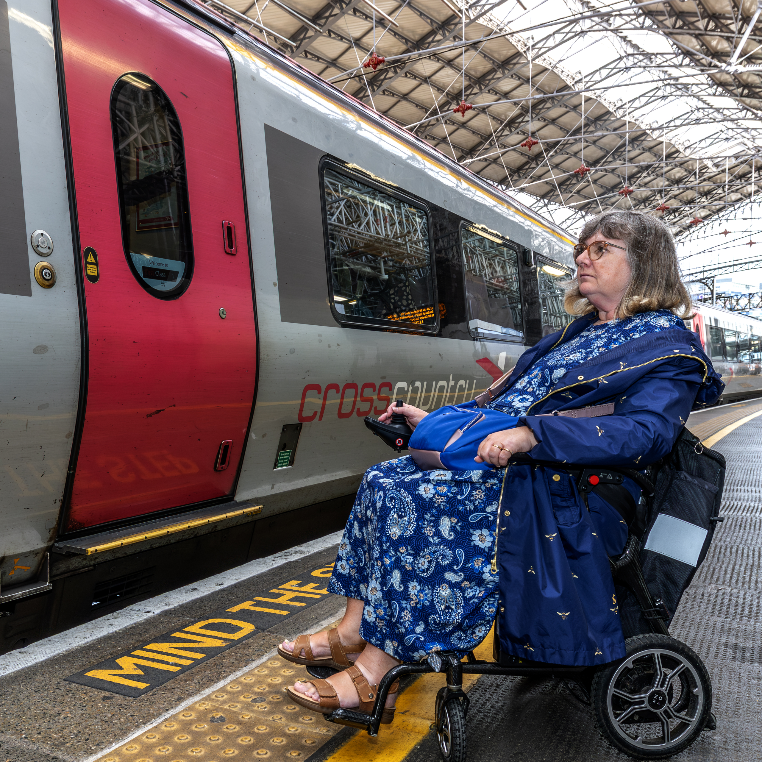 Female wheelchair user in blue dress waits on train platform behind tactile paving. Train, tracks and station to her left in the image.