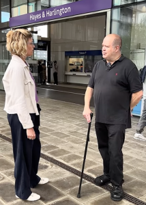 Two people standing outside a London tube station entrance, in conversation. One is a man in dark top and trousers holding a mobility aid. The other is woman in a white jacket and dark trousers.