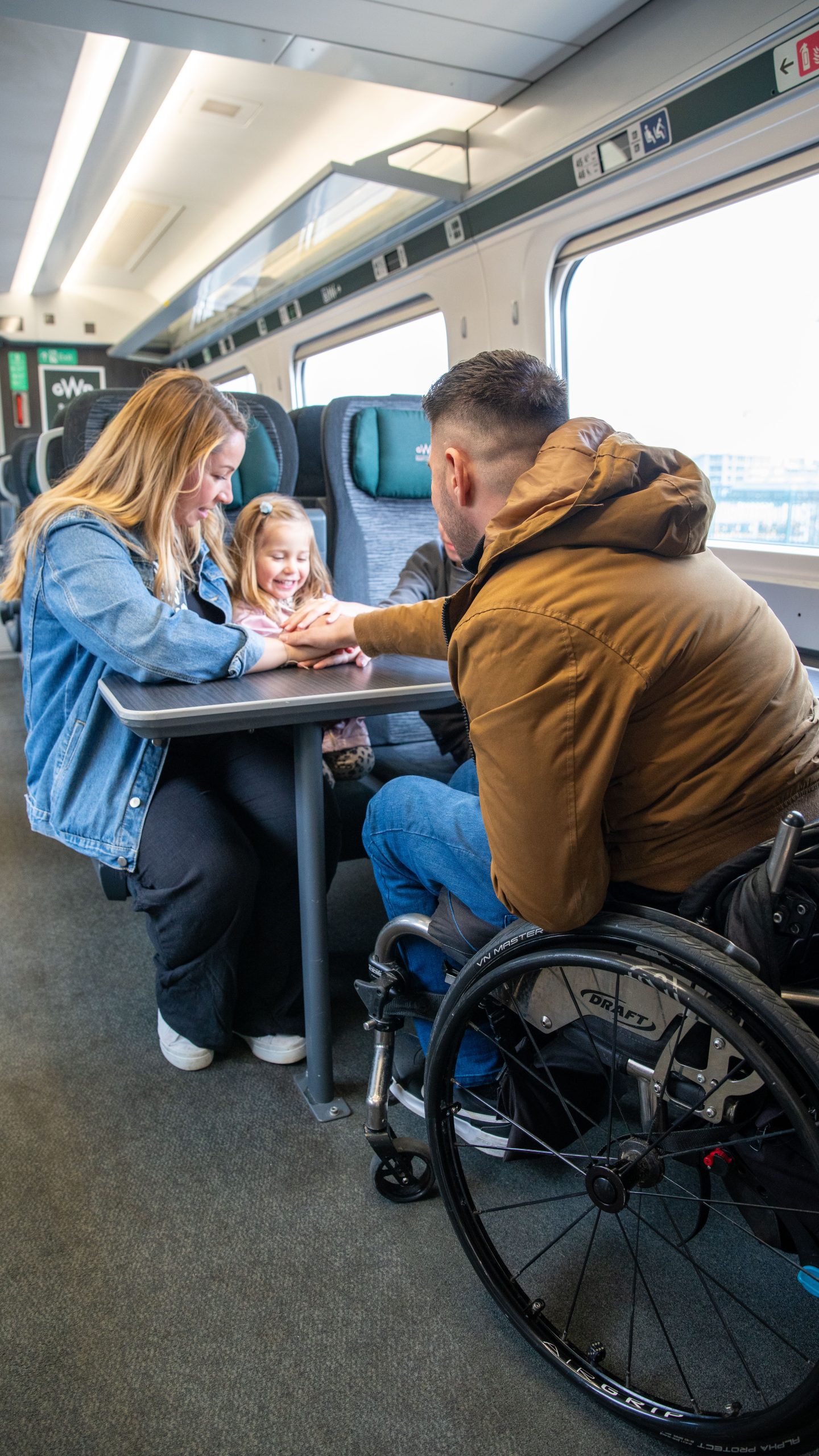 Male wheelchair user has his back to the camera as he faces his family (female partner and female child) in train carriage seating next to window.