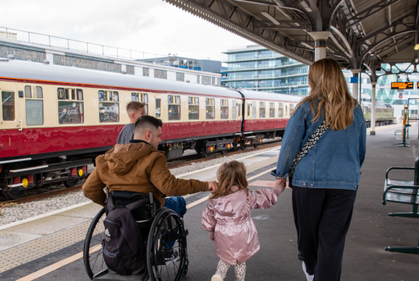 A family walks along a railway station platform. An adult wheelchair user moves alongside a woman and a young child holding hands, with a train stopped beside them on a train platform. The ncat logo and “three years” branding appear on the top of the image.