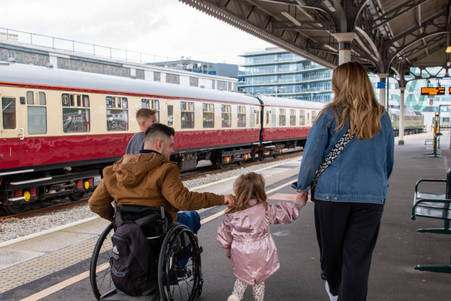 A family walks along a railway station platform. An adult wheelchair user moves alongside a woman and a young child holding hands, with a train stopped beside them on a train platform. The ncat logo and “three years” branding appear on the top of the image.
