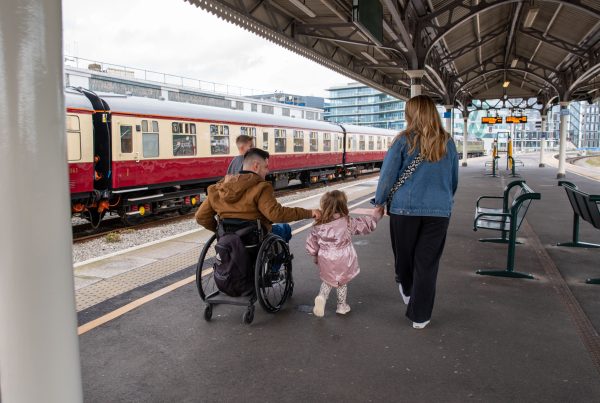 A family walks along a railway station platform. An adult wheelchair user moves alongside a woman and a young child holding hands, with a train stopped beside them on a train platform.