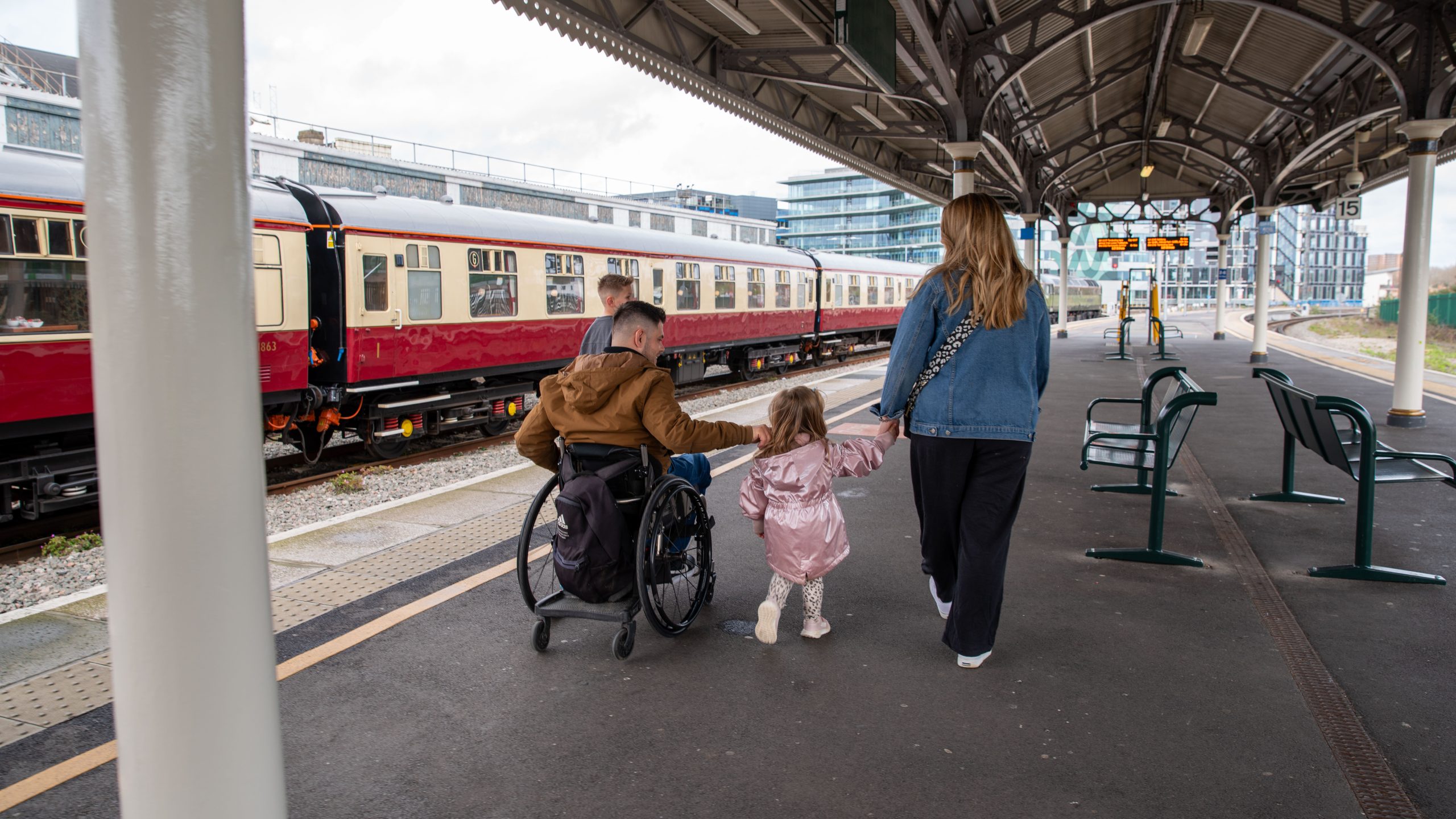 A family walks along a railway station platform. An adult wheelchair user moves alongside a woman and a young child holding hands, with a train stopped beside them on a train platform.