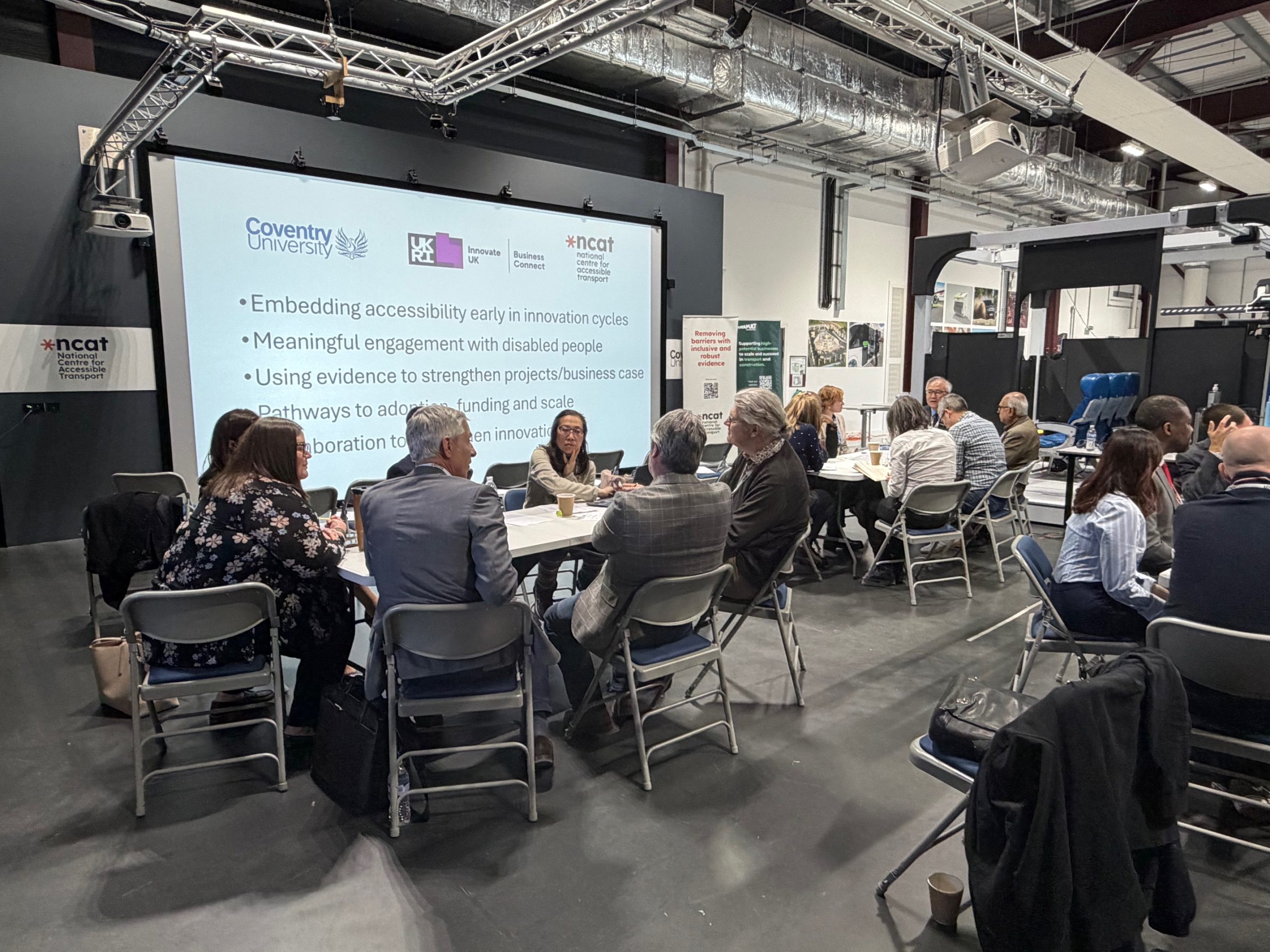 Groups of professionals seated at round tables in a modern workshop space, taking part in discussions on accessible transport innovation.