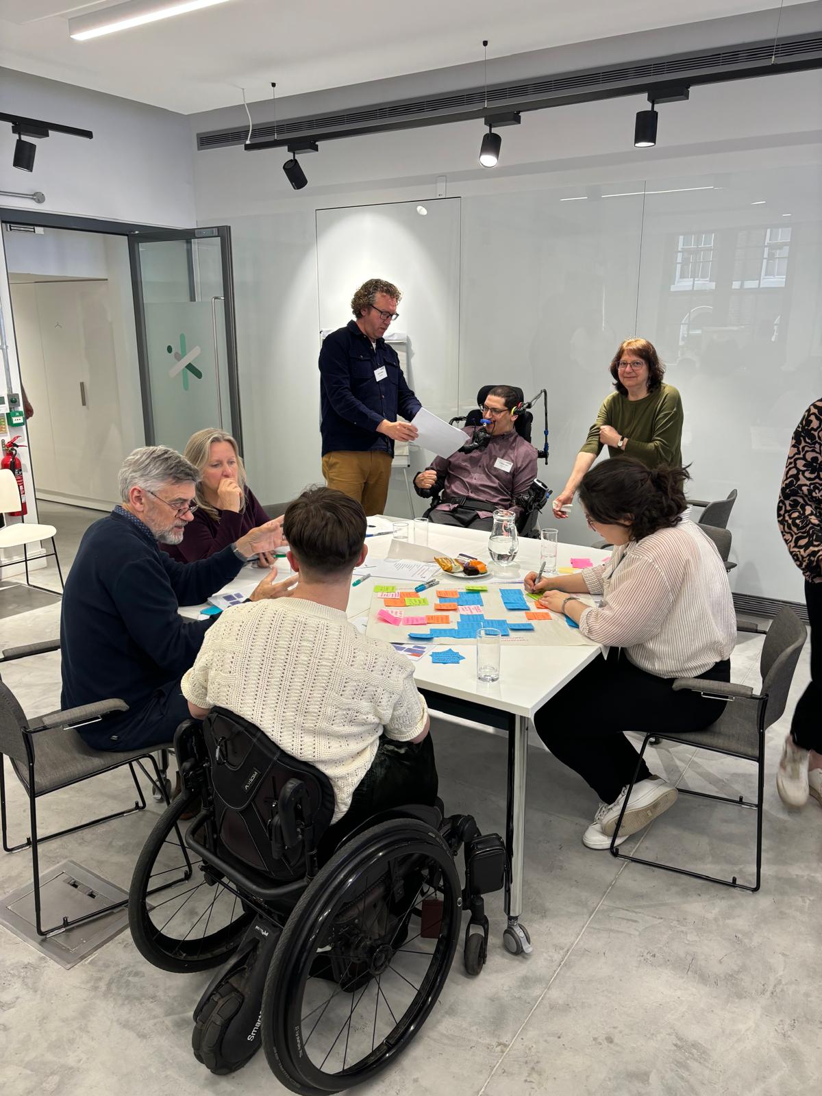 A group of people are sitting and standing round a table covered in sticky notes and pens in a plain white meeting room.