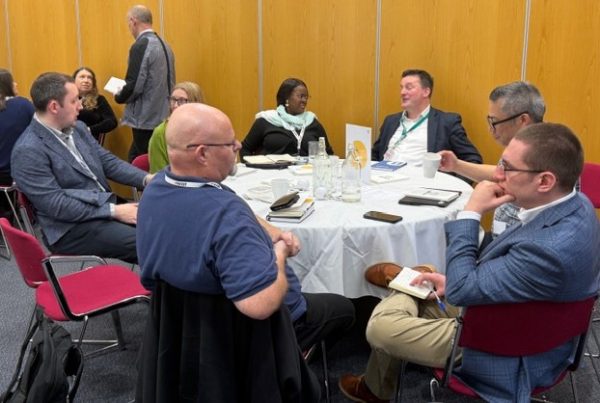Six people sitting at round table in a meeting room with brown walls having a discussion.
