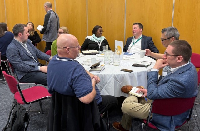 Six people sitting at round table in a meeting room with brown walls having a discussion.