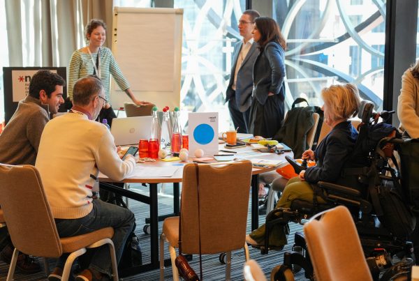 ncat staff and stakeholders sit and stand around a table in a meeting room with a mini whiteboard and window in the background.
