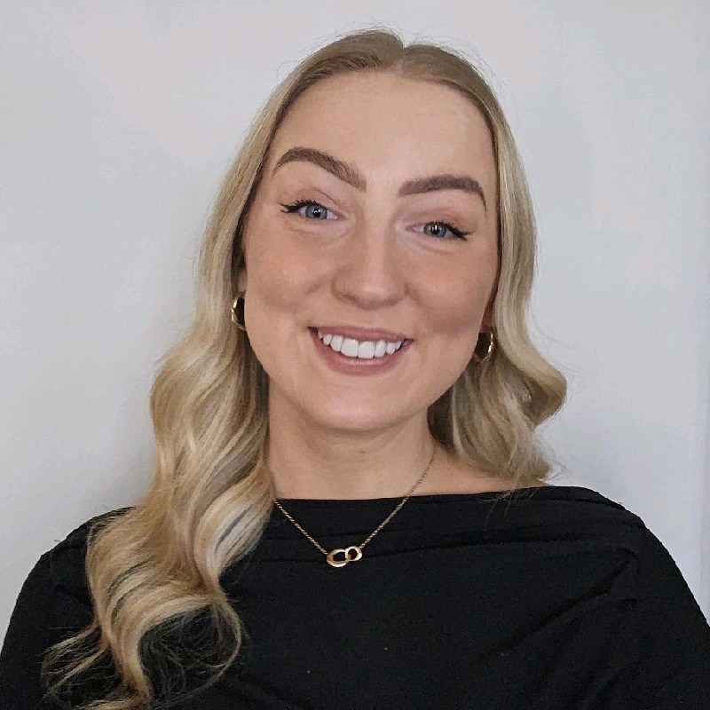 a photo of a white woman with long blond hair, wearing a black top with a gold necklace, smiling