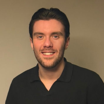 A photo of a white male with short brown hair and beard, wearing a black top, smiling