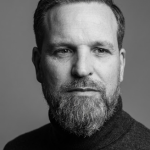 A black and white photo of a white male with moustache and a beard, wearing a dark top - looking into the camera